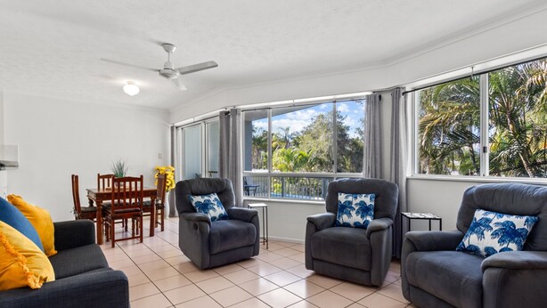 A spacious living area with a ceiling fan, two armchairs with blue pillows, a sofa with yellow pillows, a dining table with chairs, and large windows offering a view of palm trees. - Spacious Esplanade Apartment With Rooftop Access (Hervey Bay)