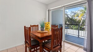 A dining area with a wooden table and chairs, a potted plant, and a view of the outdoors through a sliding glass door. - Spacious Esplanade Apartment With Rooftop Access (Hervey Bay)