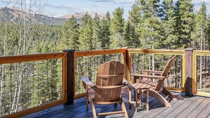 Una terraza de madera con sillas Adirondack y una mesa, con vista a un bosque y montañas.