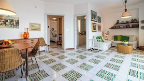 A spacious living area with a dining table, chairs, and a sofa. The floor has a patterned tile design. There are shelves with books and decorative items. - Sea Escape - Apartment by the Sea (Giardini Naxos)