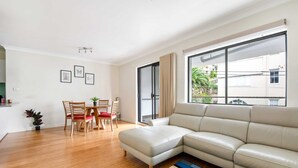 A modern living room with a beige leather sofa, wooden dining table with chairs, and a large window with a view of greenery. - Lovely 2-bed With Balcony (Sydney)