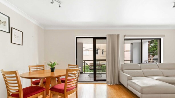 A modern living room with a wooden dining table, red cushioned chairs, a grey sofa, and a sliding glass door leading to a balcony. - Lovely 2-bed With Balcony (Sydney)