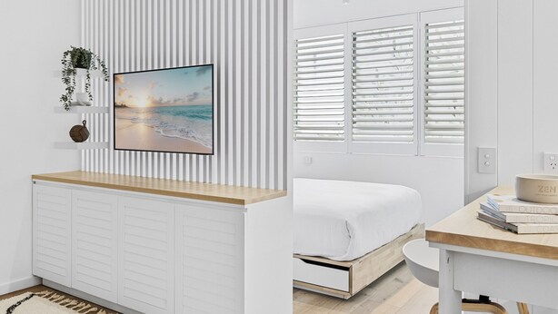 A modern bedroom with a bed, a bedside table, a wall-mounted TV, and a window with blinds. - Studio4: Bondi Chic Interiors With Rooftop Pool (Sydney)