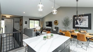 Uma cozinha moderna com bancada de mármore, uma sala de jantar com mesa e cadeiras de madeira, e uma sala de estar com sofá e televisão.
