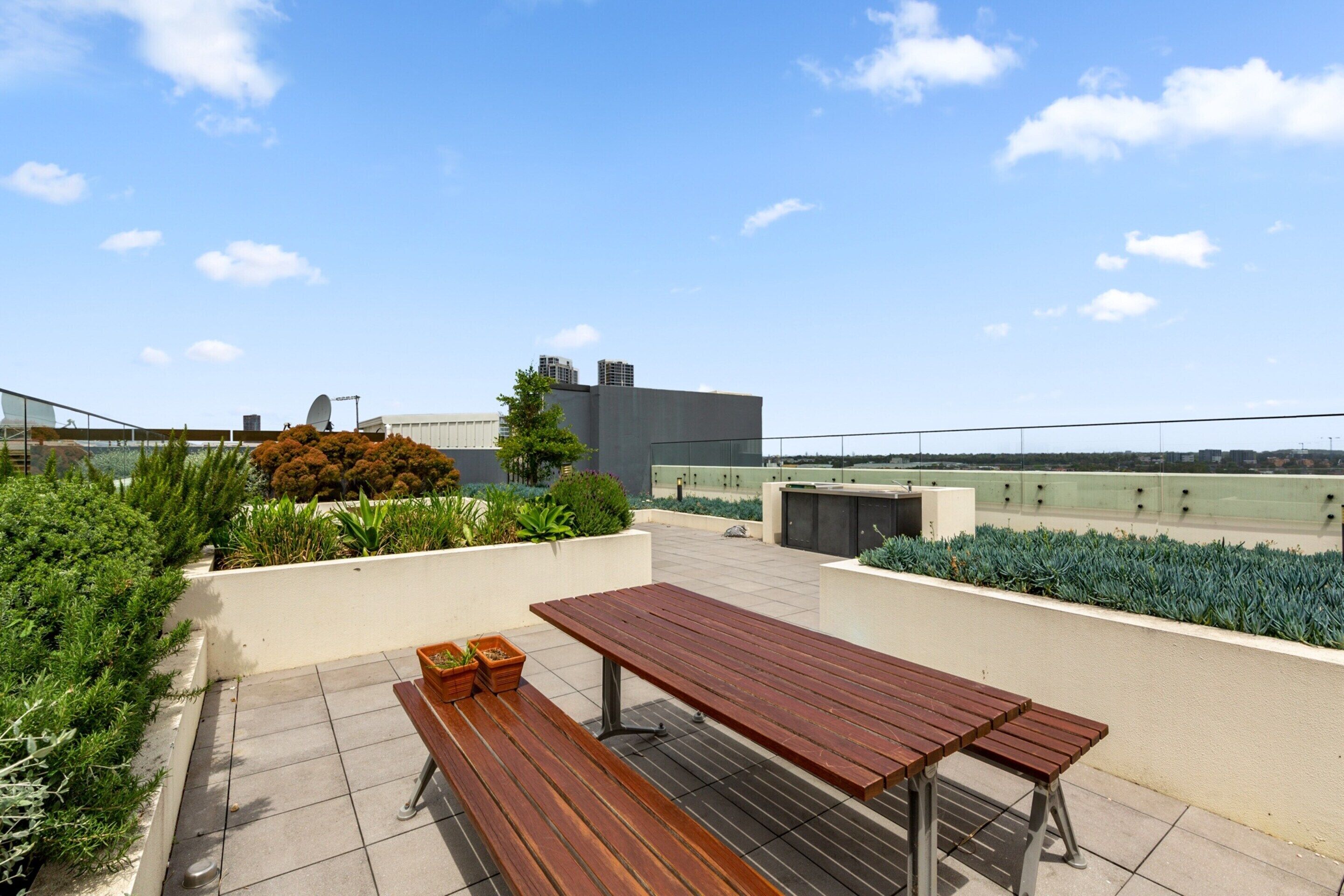 A rooftop terrace with a wooden table and benches, surrounded by greenery and a clear sky.