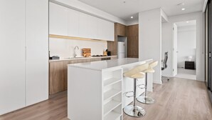 A modern kitchen with white cabinetry, a central island with bar stools, and a wooden accent wall. - Luxe 2-bed: Modern Interiors, Balcony & BBQ Area (Sydney)