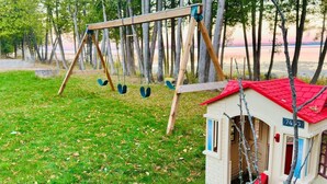 A small wooden swing set with four seats, a small red-roofed structure with a mailbox, and a grassy area with trees in the background. - Lakefront 2-BR Cottage Retreat in Kagawong - Lilly Lakehouse (Billings)
