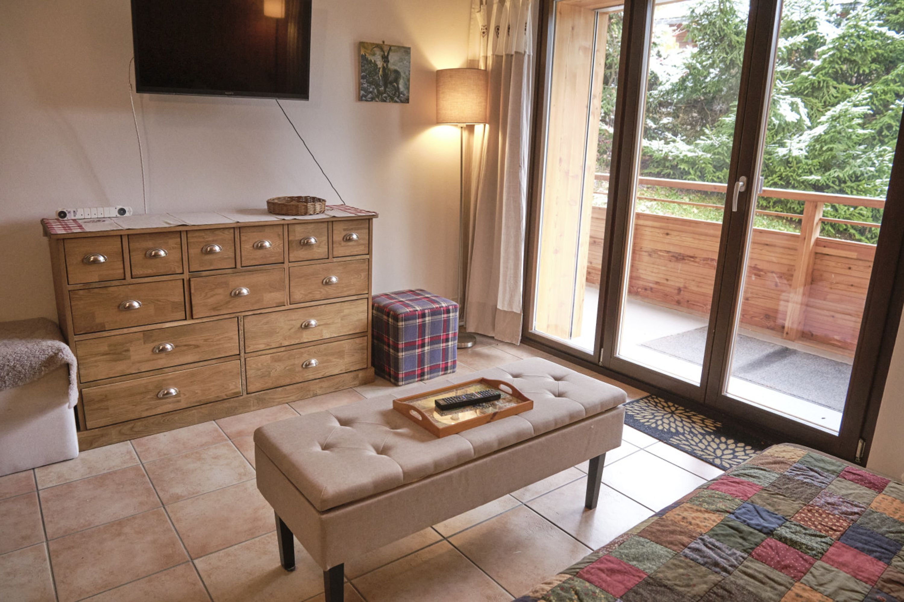 A bedroom with a wooden dresser, a bench with a tray, a television, and a view of a wooden deck and greenery outside.