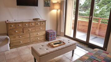 A bedroom with a wooden dresser, a bench with a tray, a television, and a view of a wooden deck and greenery outside.