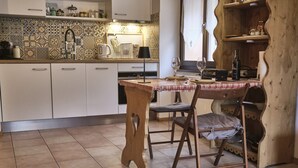 A kitchen with white cabinets, a tiled backsplash, a wooden table with a checkered tablecloth, and a wooden chair. - Studio Pierre a Voir 4 (Riddes)