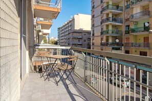 A balcony with a table and chairs, overlooking a street with parked cars and multi-story buildings.