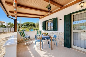En overdækket terrasse med et bord og stole, en hængende plante og en stenvæg.