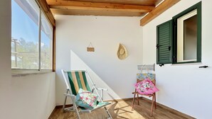 A room with a wooden ceiling, a window with green shutters, a green and white striped chair, a pink chair with floral upholstery, and a wall-mounted clock.