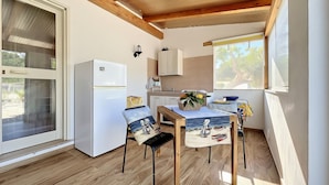 A compact kitchen with a refrigerator, wooden ceiling beams, a dining table with a placemat, and a window with a view of trees.