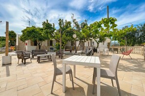 Un patio al aire libre con una mesa blanca y sillas de mimbre, rodeado de árboles y un cielo despejado.