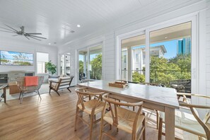 A spacious dining area with a large table and wicker chairs, a ceiling fan, and large windows offering views of the surrounding area.