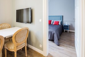 Quarto com televisão na parede, cama com cabeceira azul e mesa de cabeceira com abajur.