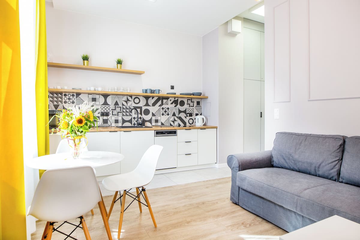 Una cucina moderna con mobili bianchi, un piano di lavoro in legno e una schiena di ripiano con motivi geometrici. C'è un tavolo da pranzo con sedie bianche e un vaso di fiori. In zona soggiorno si trova un divano grigio.
