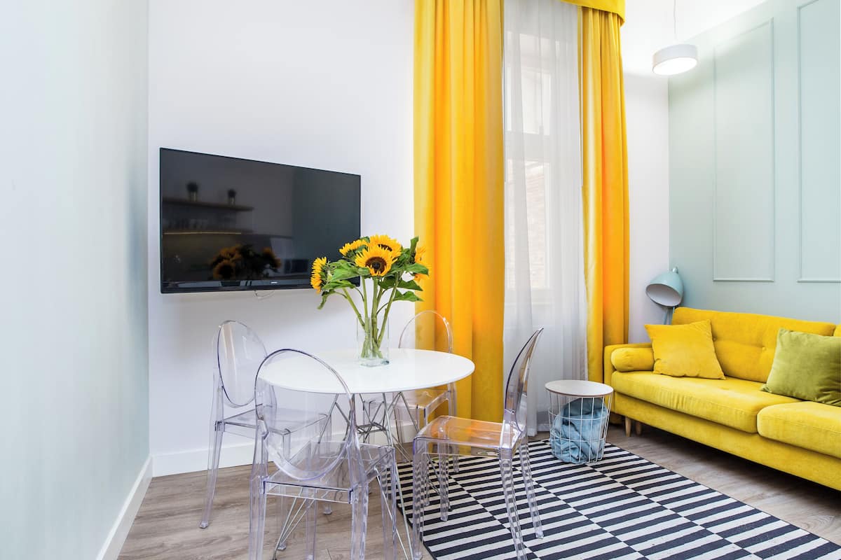Un soggiorno moderno con un divano giallo, un tavolino rotondo con sedie trasparenti, una TV a parete e un vaso con girasoli.