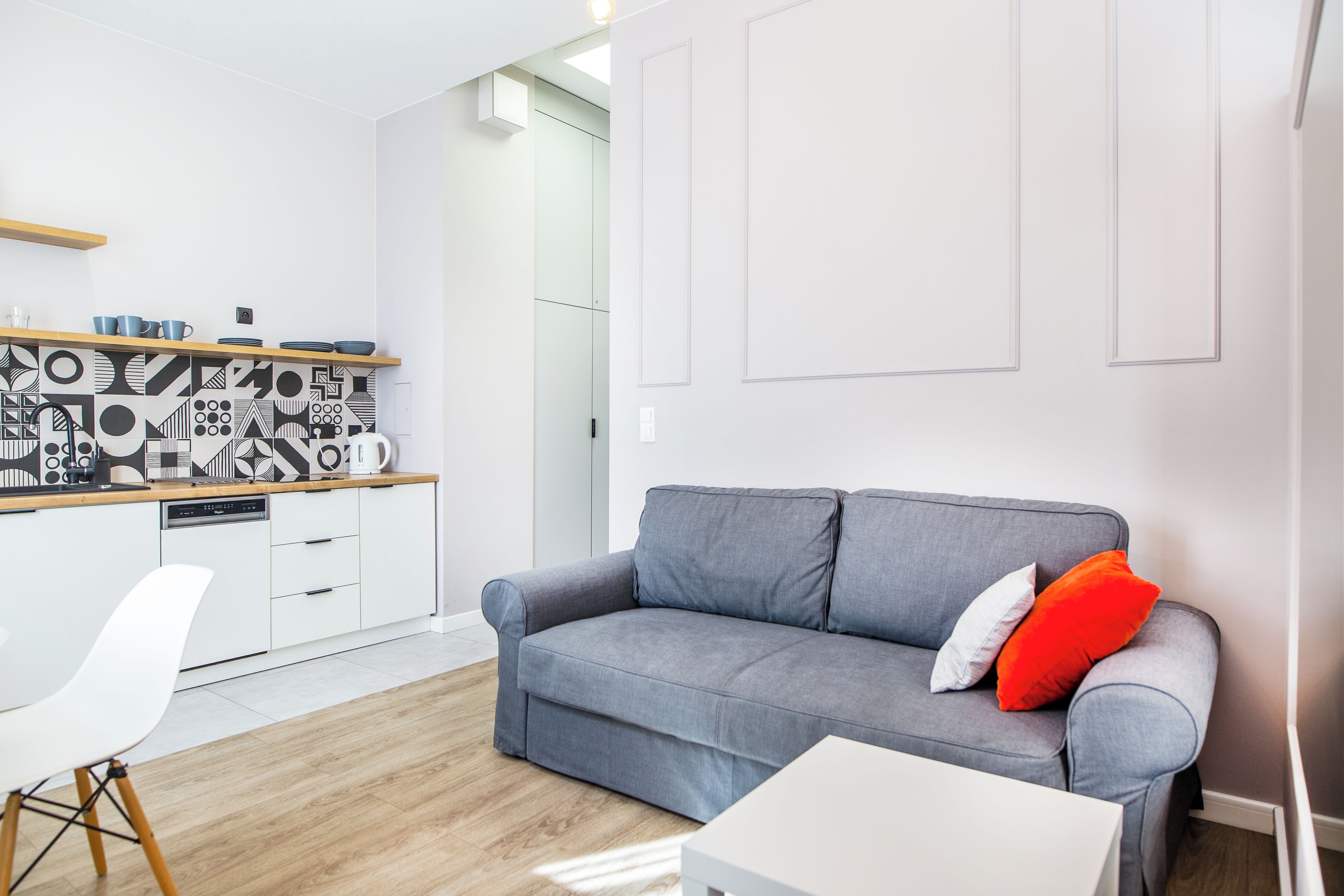 A modern living room with a grey sofa, a white coffee table, and a white chair. There is a kitchen area with white cabinets and a patterned backsplash.