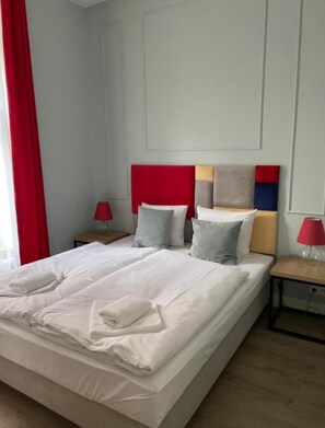 Un lit bien fait, avec du linge de lit blanc et deux oreillers, une tête de lit rouge et une table de chevet avec une lampe.