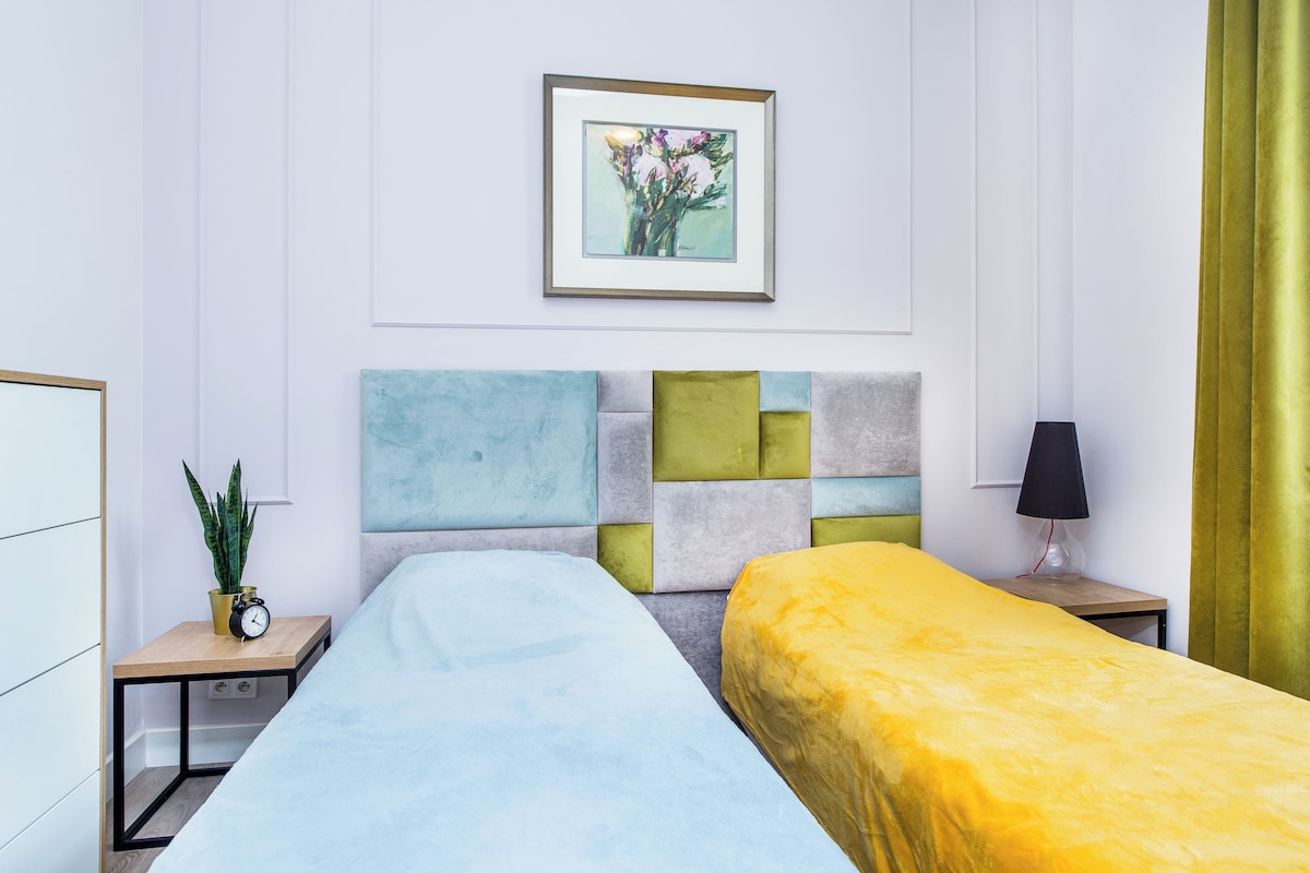 Une chambre d’hôtel avec deux lits, une table de chevet avec une horloge et une plante, un tableau encadré au mur et une lampe sur la table de chevet.