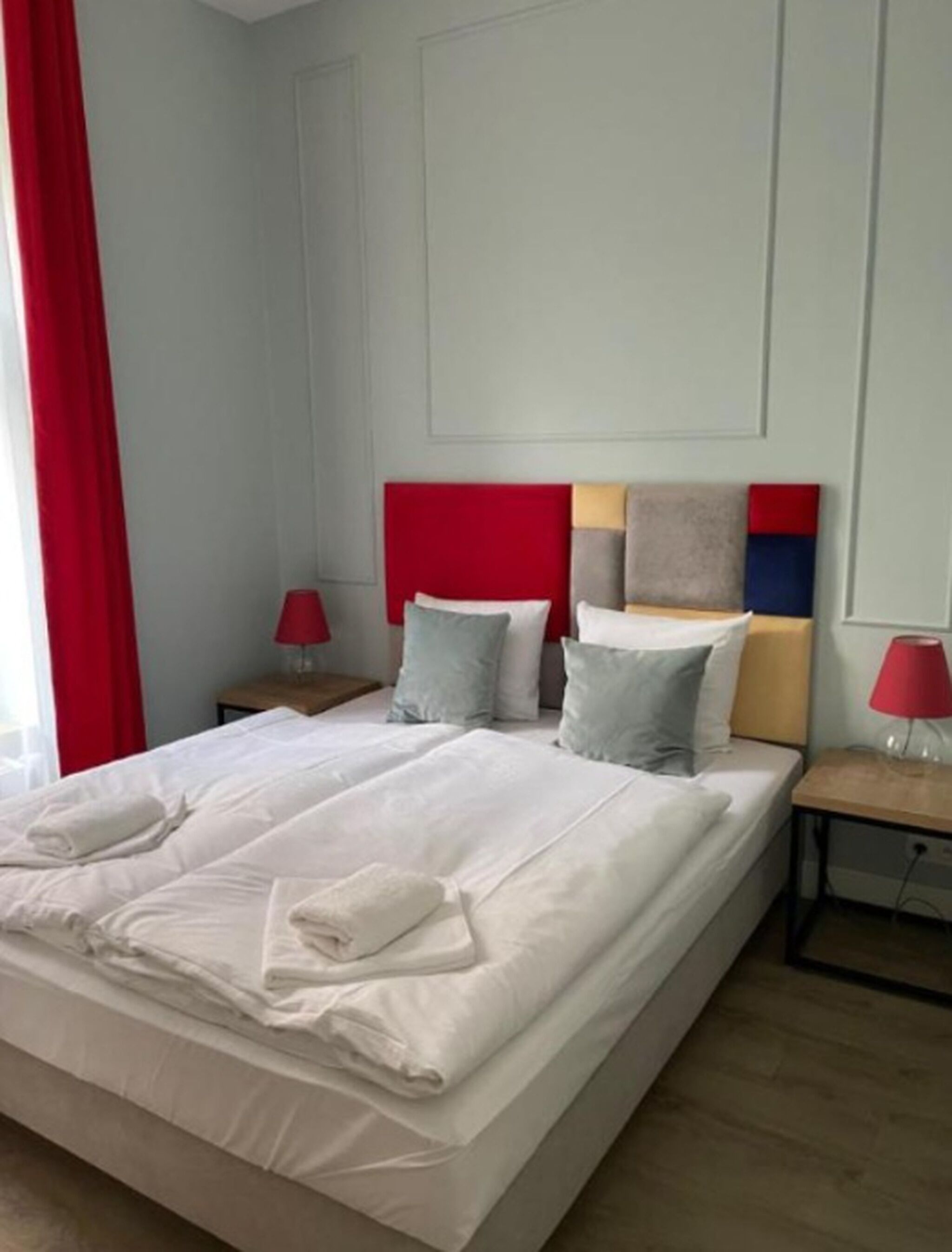 A neatly made bed with white linens and two pillows, a red headboard, and a bedside table with a lamp.