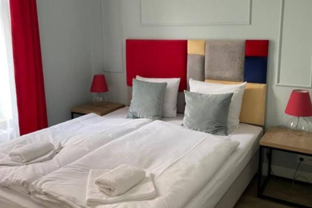 Un letto rifatto con lenzuola bianche e due cuscini, una testiera rossa e un comodino con una lampada.