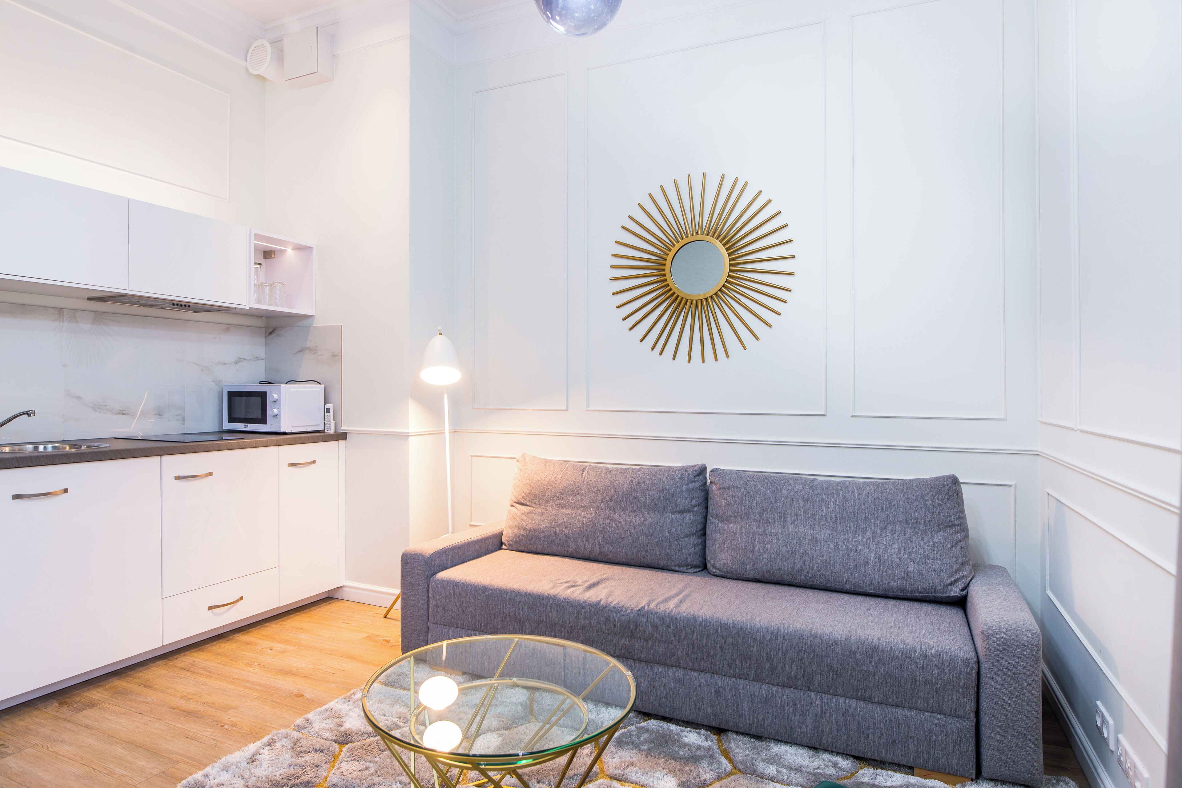 Een moderne keuken met witte kasten, een grijze bank en een spiegel in zonnegloed op de muur.