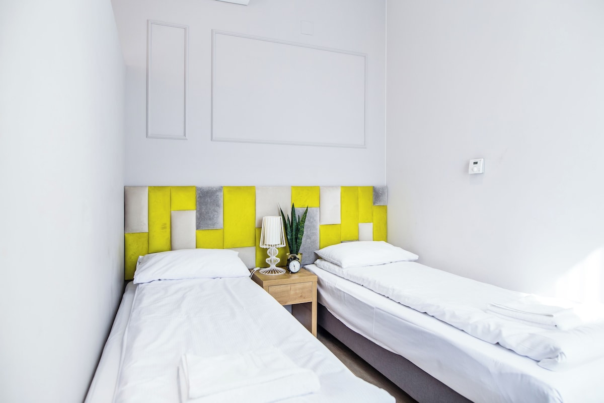 Une chambre d’hôtel avec deux lits simples, une table de chevet en bois avec une lampe, un petit réveil et une plante en pot.