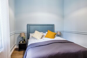 A neatly made bed with a blue headboard, two pillows, a dark blanket, and a nightstand with a lamp and a clock.