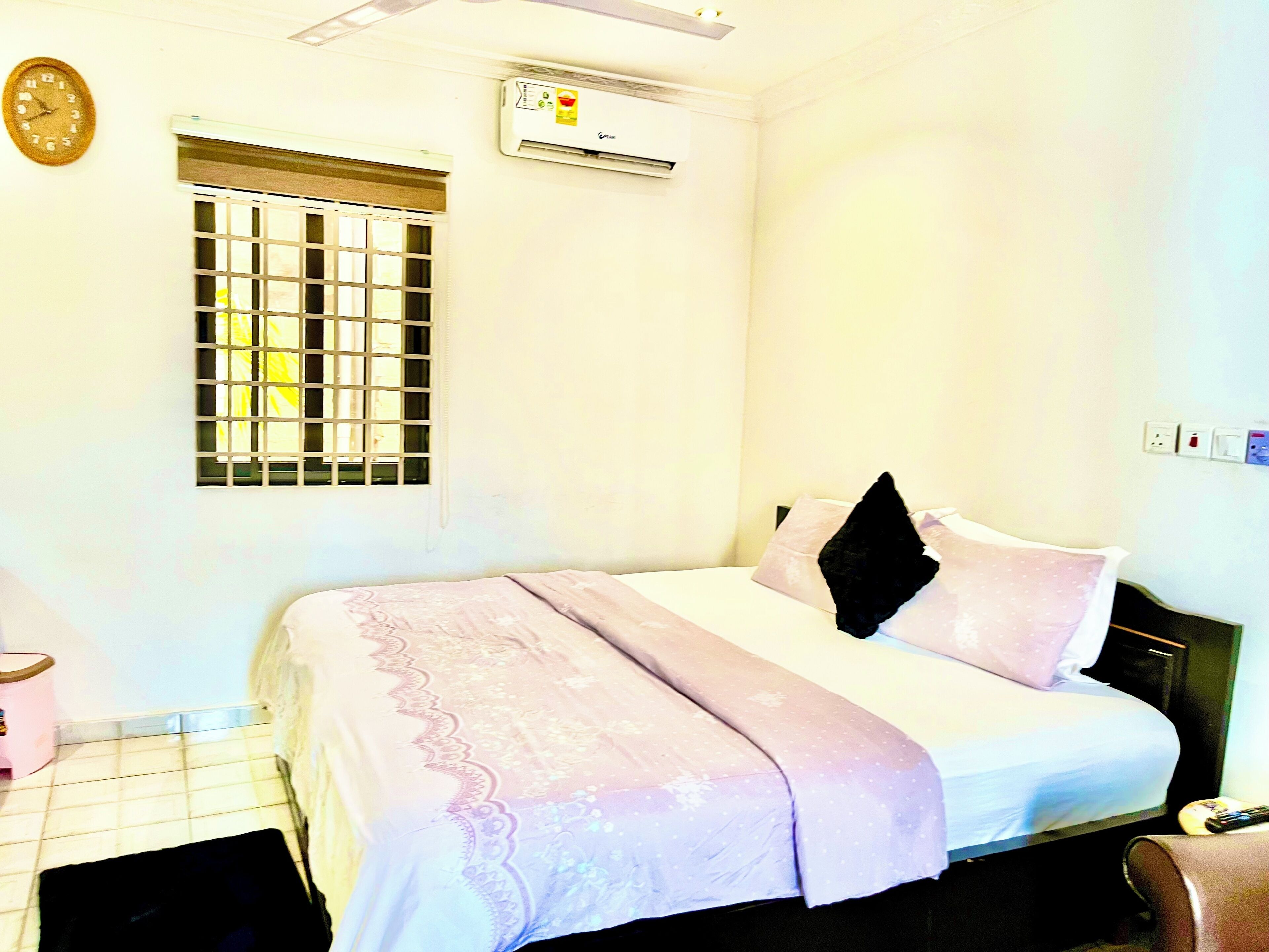 A hotel room with a bed, a window with bars, an air conditioning unit, a clock, and a power outlet.