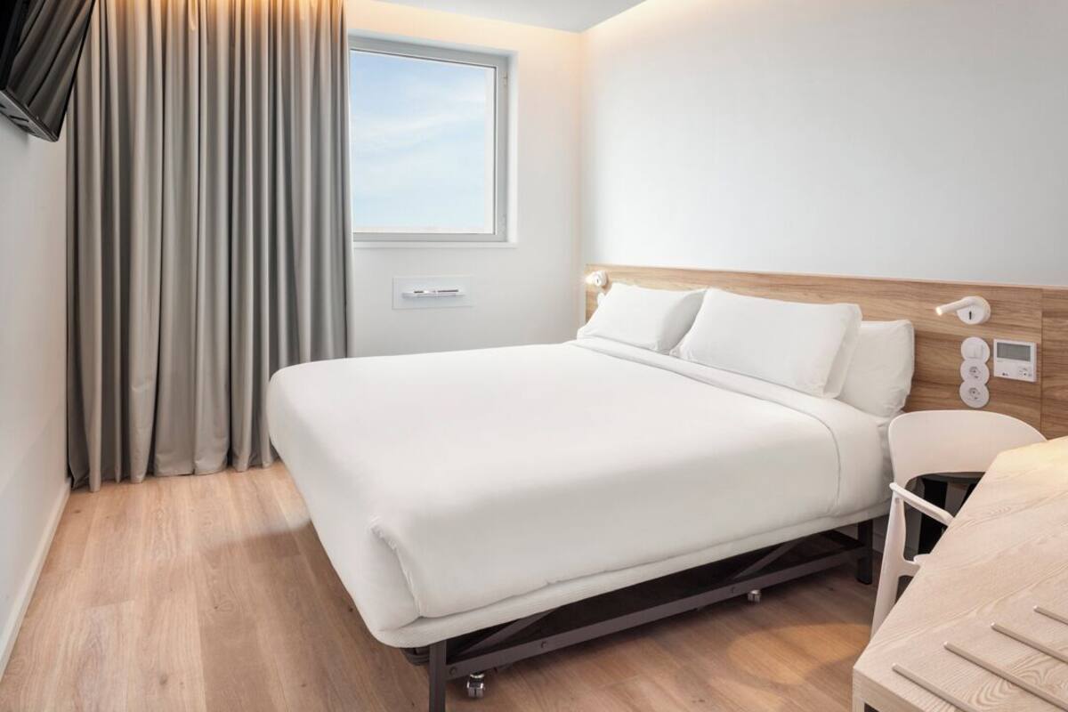Une chambre d’hôtel moderne avec un grand lit, une table de chevet, une lampe et une fenêtre avec des rideaux.
