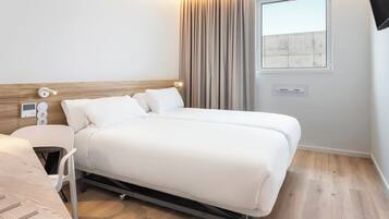Quarto de hotel moderno com cabeceira de madeira, cama com roupa de cama branca, mesa de cabeceira, cadeira, janela com cortinas e vista para um edifício.