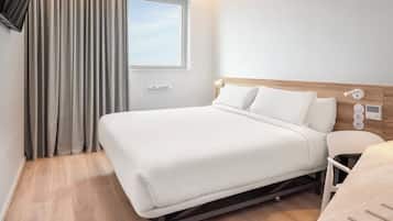 A modern hotel room with a bed, a bedside table, a lamp, a window with curtains, and a wooden floor.