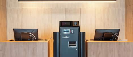 A modern hotel room with a wooden wall paneling, a central black minibar, and two laptops on wooden desks.