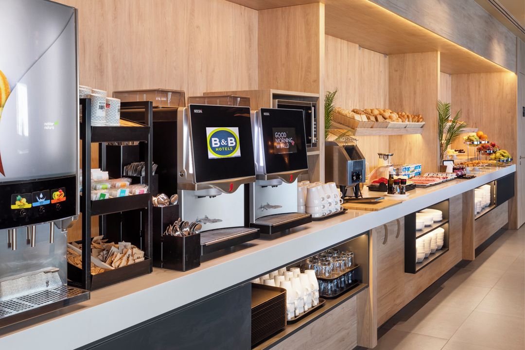 A B&B hotel breakfast area with coffee machines, a fruit display, and a variety of breakfast items.
