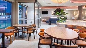 A modern hotel lobby with round wooden tables and orange chairs, a large potted plant, and a bar area. - B&B Hotel Vila Do Conde (Vila do Conde)