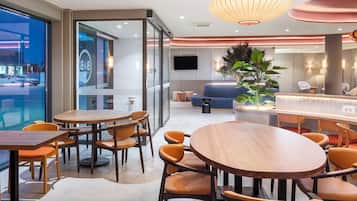 A modern hotel lobby with round wooden tables and orange chairs, a large potted plant, and a bar area.