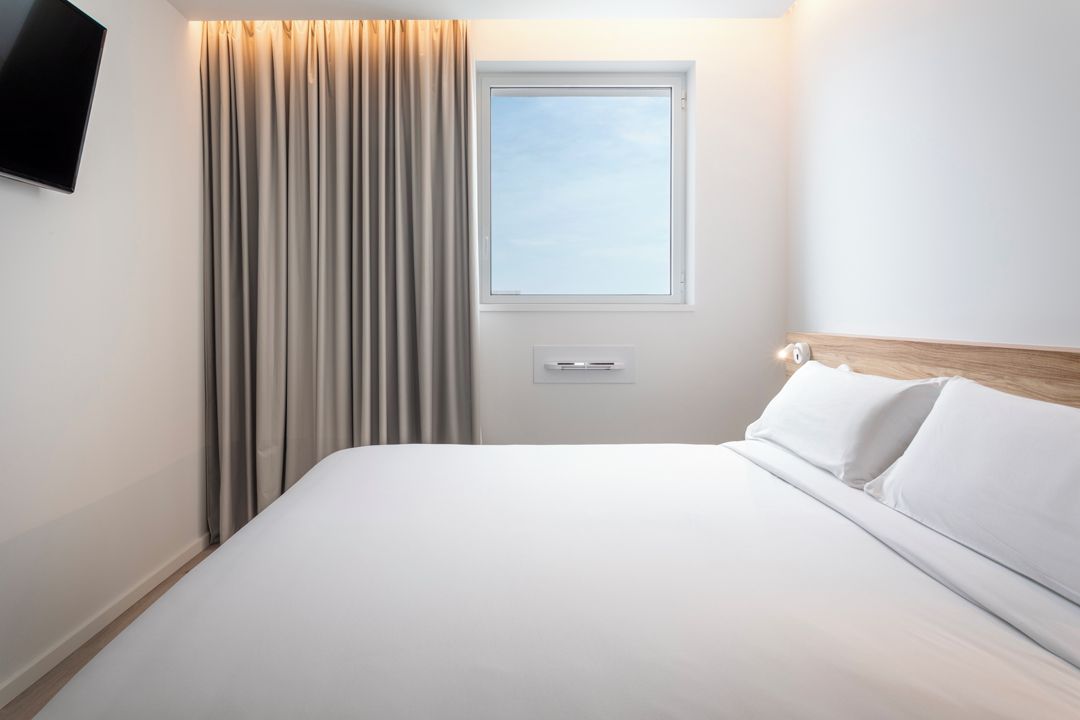 Quarto de hotel com uma cama, janela, cortinas, televisão fixada na parede e uma porta.