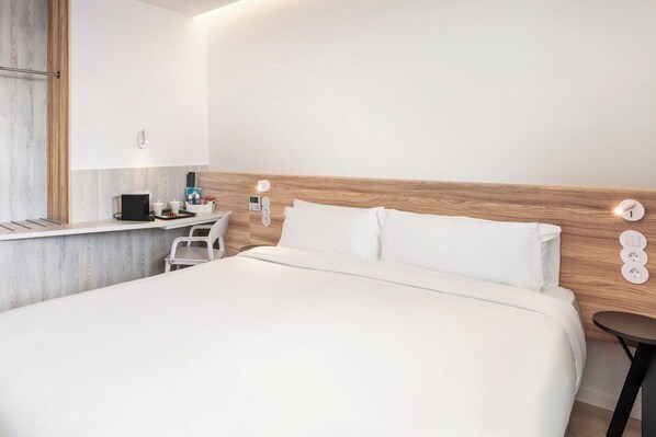 Une chambre d’hôtel moderne avec une tête de lit en bois, un lit recouvert de draps blancs, une petite table de chevet avec une lampe et une prise électrique fixée au mur.