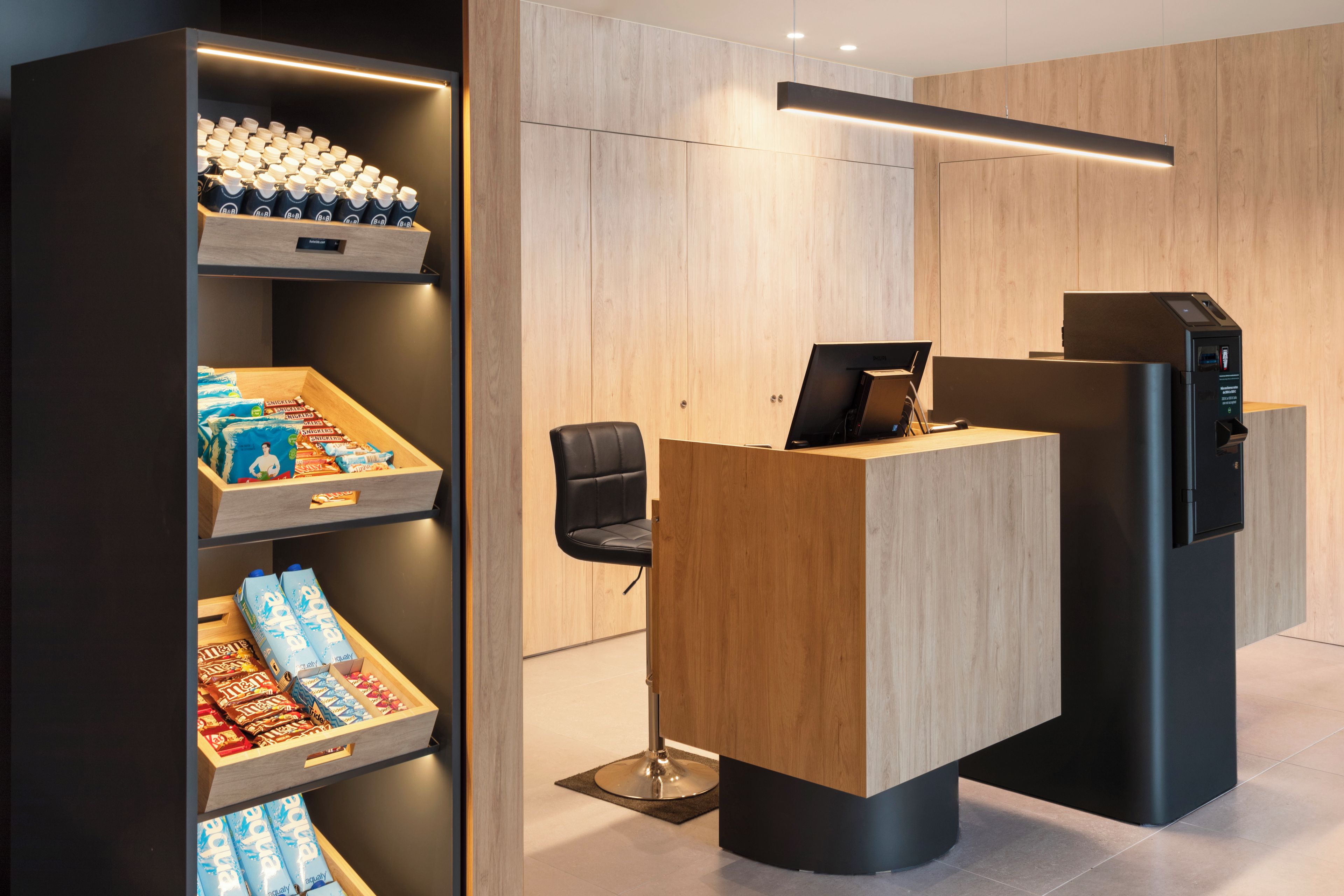 Un espace d’accueil avec un bureau en bois, une chaise noire et une étagère contenant des bouteilles d’eau et des en-cas.