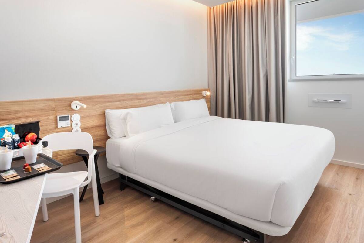 Une chambre d’hôtel comprenant un lit, un bureau avec une chaise, une petite table avec quelques objets, une fenêtre avec des rideaux et un panneau de commande fixé au mur.