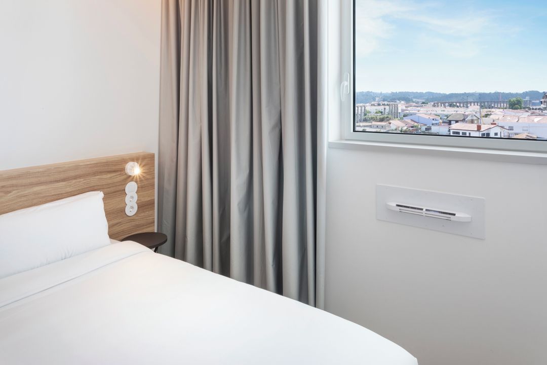 Habitación de hotel con cama, ventana con vista a la ciudad, aire acondicionado de pared y lámpara.