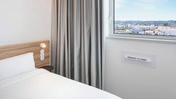 Habitación de hotel con cama, ventana con vista a la ciudad, aire acondicionado de pared y lámpara.