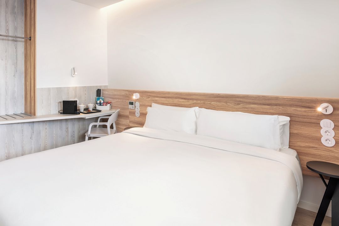 Een moderne hotelkamer met een houten hoofdeinde, een bed met witte lakens, een klein nachtkastje met een lamp en een wandcontactdoos.