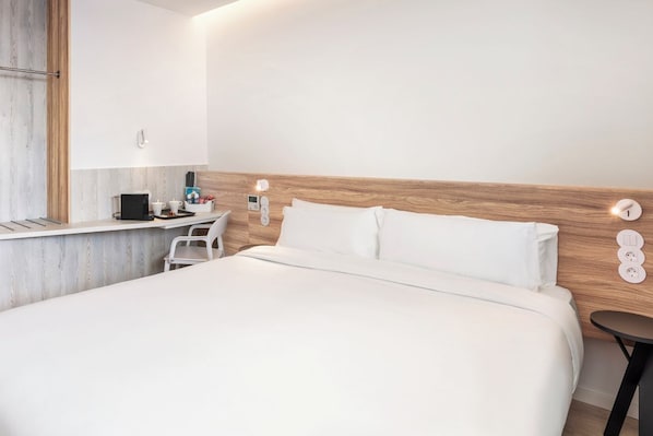 A modern hotel room with a wooden headboard, a bed with white linens, a small bedside table with a lamp, and a wall-mounted power outlet. - B&B Hotel Vila Do Conde (Vila do Conde)