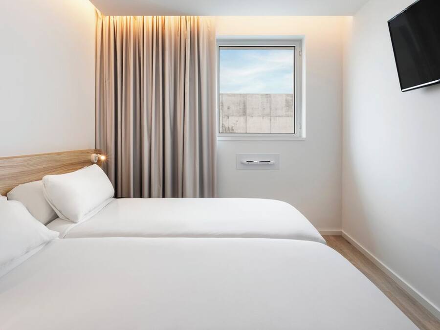 A modern hotel room with a bed, a window with curtains, a mounted TV, and a wall-mounted air conditioning unit.