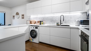 A modern kitchen with white cabinetry, a built-in washing machine, a microwave, and a sink with a hexagonal backsplash. - Riverside 2-bed Apartment With Balcony & Parking (Melbourne)