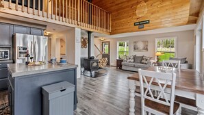A spacious living area with a kitchen, dining table, and a loft with a wooden railing. - S Diamond S Retreat | Huge Backyard & Hot Tub – 2 Miles to Town! (Rangeley)